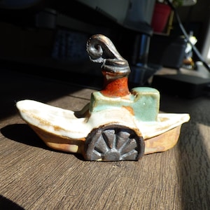 May include: A handcrafted ceramic boat sculpture with a whimsical design. The boat features a white hull, a green cabin, and a red and brown smokestack. The front of the boat has a dark brown wheel design.