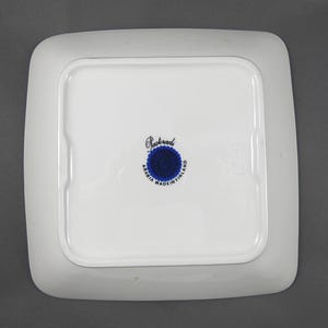 K&ouml;nnte beinhalten: Ein quadratischer, wei&szlig;er Keramikteller mit abgerundeten Ecken. Die Unterseite zeigt ein zentrales blau-wei&szlig;es Emblem mit dem Text "Arabia Made in Finland" und dem Namen "Pasteraali". Der Teller hat einen leicht erh&ouml;hten Rand.