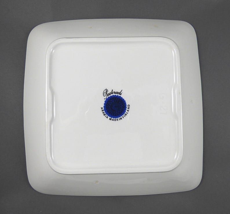 K&ouml;nnte beinhalten: Ein quadratischer, wei&szlig;er Keramikteller mit abgerundeten Ecken. Die Unterseite zeigt ein zentrales blau-wei&szlig;es Emblem mit dem Text "Arabia Made in Finland" und dem Namen "Pasteraali". Der Teller hat einen leicht erh&ouml;hten Rand.