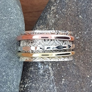 May include: A wide spinner ring featuring a central band with an intricate etched swirl pattern, flanked by three smooth, thin rotating bands in copper, silver, and gold tones. The ring is constructed from multiple metal layers with contrasting textures.