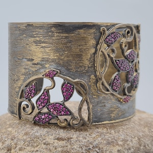 Puede incluir: Un brazalete ancho con un acabado metálico oscuro y texturizado que presenta dos secciones recortadas. Estas áreas están decoradas con motivos de enredaderas metálicas entrelazadas, incrustadas con numerosas piedras rosas.