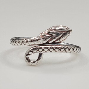 Puede incluir: Un anillo de metal plateado con forma de serpiente enroscada. La banda presenta un patrón de escamas detallado y texturizado en toda su longitud, con la cabeza de la serpiente en un extremo y una cola curva formando el otro extremo del anillo.