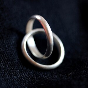 May include: Two interlocking silver rings with a smooth, matte finish. The bands are circular and feature a consistent, rounded profile. The metal has a soft, reflective quality that catches the light, highlighting the simple, clean design of the jewelry.