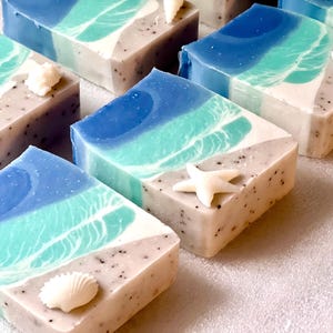 May include: Artisan soap bars with a beach theme. Each bar features layers of blue and teal, resembling ocean waves, and a sandy base with small dark specks. Decorative seashells and starfish are placed on top of the bars.