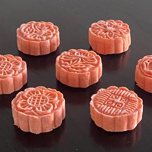 May include: Seven round, reddish-brown mooncakes with intricate floral and geometric designs on top. The cakes are arranged on a dark surface, showcasing their detailed patterns and textures.