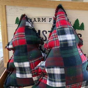 May include: Two Christmas tree-shaped pillows with a patchwork plaid design in red, green, white, and navy blue. The pillows have a frayed edge and sit in front of a wooden sign that reads "FARM FRESH".