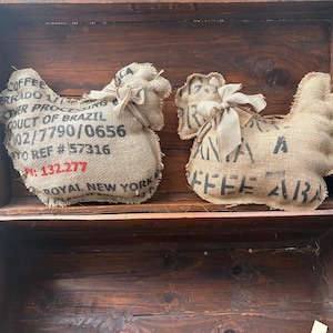 May include: Two burlap chicken-shaped decorations with text from coffee bean sacks. The chickens are tied with twine bows and rest inside a wooden box. The text includes "COFFEE ERRADO" and "ROYAL NEW YORK".