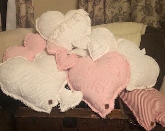 May include: A collection of heart-shaped pillows in white and pink with a textured, quilted fabric. The pillows have a frayed edge and are arranged on a wooden surface.