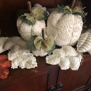 May include: A collection of white, fluffy pumpkins with burlap and green fabric accents. The pumpkins are arranged on a wooden shelf with brown and white checkered fabric leaves.