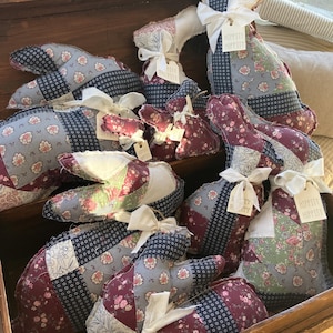 May include: A collection of patchwork fabric bunny rabbits in a wooden chest. The bunnies are in shades of blue, green, pink, and white. Each bunny has a tag that says "Hippity Hoppity".