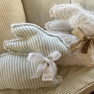 May include: Two handmade bunny pillows, one in white with a frayed texture and a brown bow, and one in green and white stripes with a white bow and a tag that says "Hippin Hoppity".