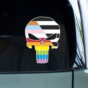 May include: A black and white skull sticker with a rainbow and gender pride flag design. The sticker is on the back window of a car.