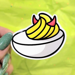 May include: A white ceramic egg-shaped dish with a yellow and red devil-themed banana filling. The dish is a fun and quirky design for serving food.
