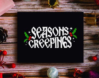 May include: A black card with white text that reads "Seasons Creepings" with a green holly sprig with red berries on each side.