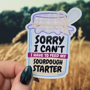 May include: A sticker featuring a jar with a purple lid and a beige string bow. The text on the sticker reads "SORRY I CAN'T I HAVE TO FEED MY SOURDOUGH STARTER". The background is blurred with green and brown tones.