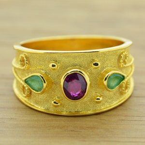 Puede incluir: Un anillo de oro con una superficie texturizada y tres piedras preciosas. La piedra central es un óvalo rosa oscuro, y las dos piedras más pequeñas son verdes y en forma de lágrima.
