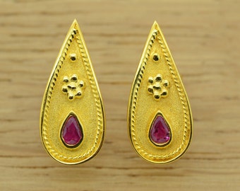 May include: A pair of gold teardrop earrings with a pink gemstone in the centre of each earring. The earrings have a detailed design with a small flower-like pattern above the gemstone.