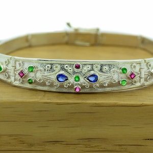 May include: A silver bracelet with an intricate design and colorful gemstones. The bracelet has a hinged clasp and is decorated with blue, green, and pink stones.