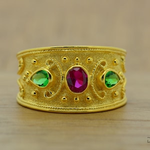 May include: A wide, gold ring with a textured surface. It features a central oval ruby and two pear-shaped green gems, with decorative details. The ring is a statement piece.