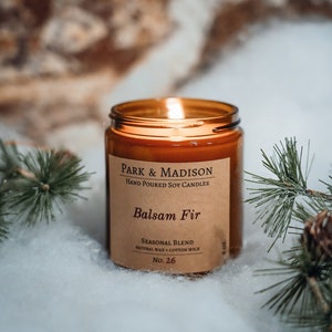 May include: A brown glass candle jar with a brown paper label that reads "PARK & MADISON HAND POURED SOY CANDLES Balsam Fir SEASONAL BLEND NATURAL WAX + COTTON WICK No. 26". The candle is lit and the flame is visible. The candle is sitting on a white surface with green pine branches.