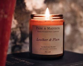 May include: A lit soy candle in a glass jar with a gold lid, labeled "Leather & Plum" by Park & Madison. The candle sits atop a dark book, with a red object in the background. The candle is a signature blend with a natural wax and cotton wick.