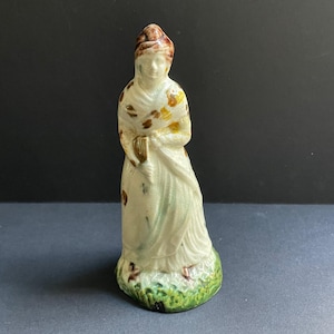 May include: A vintage ceramic figurine of a woman in a long dress and shawl, holding a book. The figurine has a white base with brown, yellow, and green accents. The base is green and resembles grass.