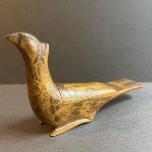 May include: A detailed wooden bird-shaped box, featuring etched patterns and a warm brown hue. The bird has a long neck and open beak, with detailed patterns on the body and wings. The box is resting on a flat surface.