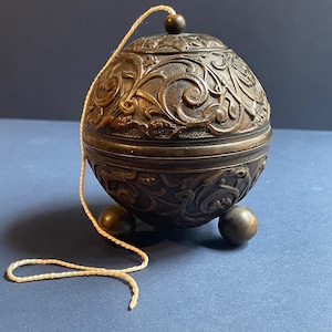 May include: A bronze-colored, spherical container with intricate floral carvings. The container rests on three small ball feet and has a matching spherical lid with a small wooden knob. A cream-colored string is attached.