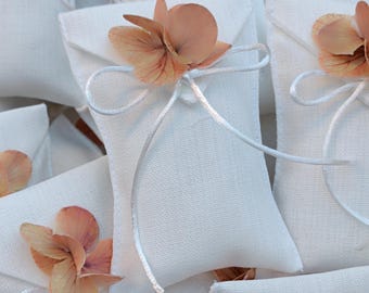 May include: A collection of small, rectangular white fabric pouches, each tied with a thin white ribbon and adorned with a single dried peach-colored hydrangea flower. The pouches have a textured surface and are arranged in a layered pile.