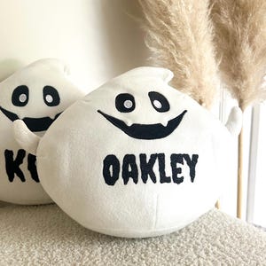 Puede incluir: Dos almohadas de felpa blancas con forma de fantasma con ojos negros, sonrisas y los nombres "K" y "OAKLEY" en letras negras. Las almohadas tienen pequeños apéndices en forma de brazo y están sobre una superficie texturizada.