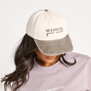 Pode incluir: Boné de beisebol creme com o texto "BE KIND TO your mind" e camiseta lilás com o texto "YOU ARE ENOUGH". O boné tem uma aba cinza. A camiseta é de cor sólida.