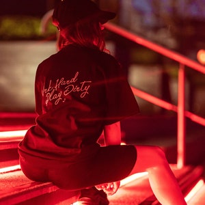May include: A person wearing a black t-shirt with the text "Work Hard Play Dirty" in pink lettering sits on a set of stairs with red LED lights.
