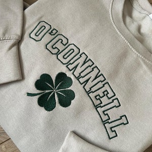 May include: A light beige sweatshirt with a green four-leaf clover and the text "O'Connell" embroidered in green.