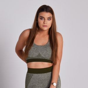 Op de afbeelding: Een vrouw draagt ​​een naadloze sportbeha in groen en grijs met bijpassende leggings. De beha heeft een donkergroene band aan de onderkant.