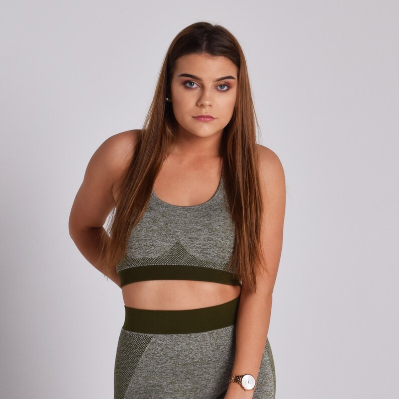 Op de afbeelding: Een vrouw draagt ​​een naadloze sportbeha in groen en grijs met bijpassende leggings. De beha heeft een donkergroene band aan de onderkant.