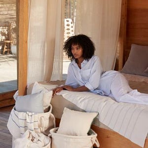 Op de afbeelding: Een slaapkamer met een bed bedekt met een lichtblauw en wit gestreept dekbed. Een persoon in lichtblauwe pyjama ligt op het bed. Drie cr&egrave;mekleurige stoffen manden bevatten kussens en een deken. De kamer heeft houten accenten en transparante gordijnen.