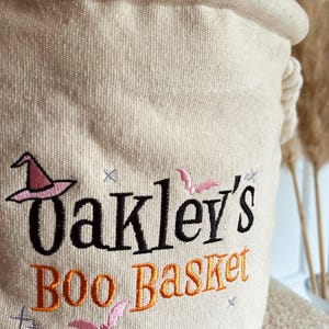 Op de afbeelding: Een beige canvas Halloween mand met de geborduurde tekst "Oakley's Boo Basket". Het ontwerp bevat een roze heksenhoed, vleermuizen en sterren in zwart en oranje draad. De mand heeft een opgerolde bovenkant en een touw handvat.