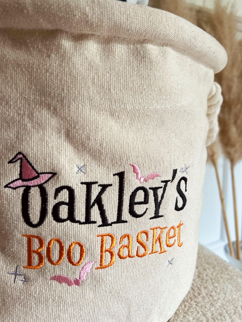 Op de afbeelding: Een beige canvas Halloween mand met de geborduurde tekst "Oakley's Boo Basket". Het ontwerp bevat een roze heksenhoed, vleermuizen en sterren in zwart en oranje draad. De mand heeft een opgerolde bovenkant en een touw handvat.