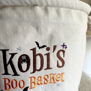 Op de afbeelding: Off-white canvas Halloween mand met de naam "Kobi's" geborduurd in bruin en de woorden "BOO Basket" in oranje. Het ontwerp bevat geborduurde vleermuizen, sterren en een spin. De mand heeft een touw handvat.