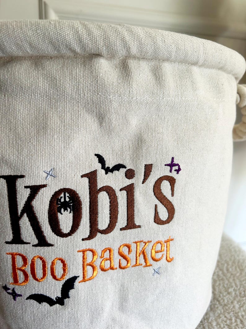 Op de afbeelding: Off-white canvas Halloween mand met de naam "Kobi's" geborduurd in bruin en de woorden "BOO Basket" in oranje. Het ontwerp bevat geborduurde vleermuizen, sterren en een spin. De mand heeft een touw handvat.