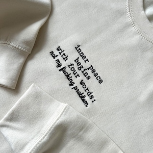 May include: Cream-colored sweatshirt with the embroidered text "inner peace begins with four words: not my f*cking problem" in black. The sweatshirt has ribbed cuffs and a crew neck.