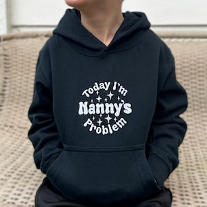 Może przedstawiać: Czarna bluza z kapturem z białym napisem "Today I'm Nanny's Problem" z gwiazdami wokół tekstu.