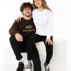 May include: A couple wearing matching sweatshirts. The woman is wearing a white sweatshirt with the text "marshmallow" in pink. The man is wearing a brown sweatshirt with the text "HOT CHOCOLATE" in gold.