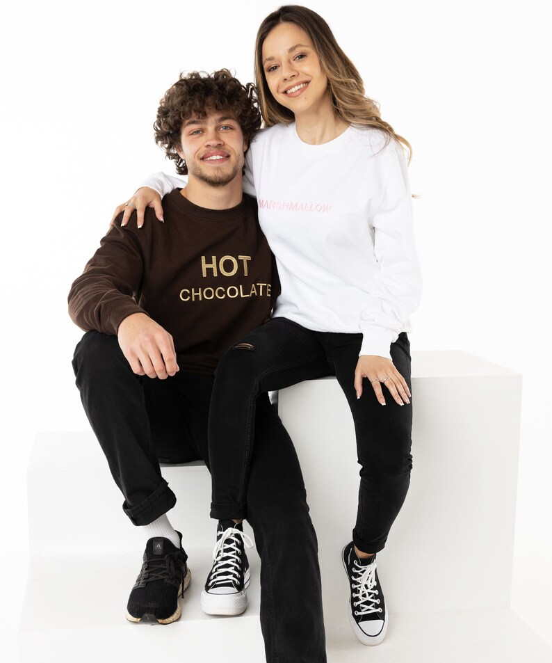 May include: A couple wearing matching sweatshirts. The woman is wearing a white sweatshirt with the text "marshmallow" in pink. The man is wearing a brown sweatshirt with the text "HOT CHOCOLATE" in gold.