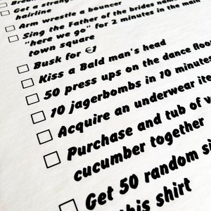 May include: A white t-shirt with a black checklist of humorous tasks, including 'Drink a pint out of a stranger's shoe' and 'Get 50 randoms of this shirt'. The text is in a bold, handwritten style.