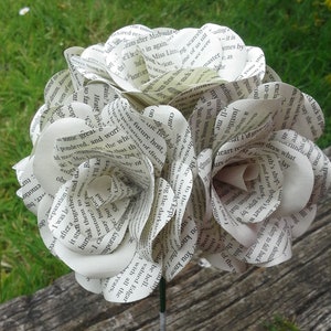 May include: A bouquet of paper flowers made from recycled book pages. The flowers are white and have a delicate, layered appearance. The bouquet is held together by a green stem.