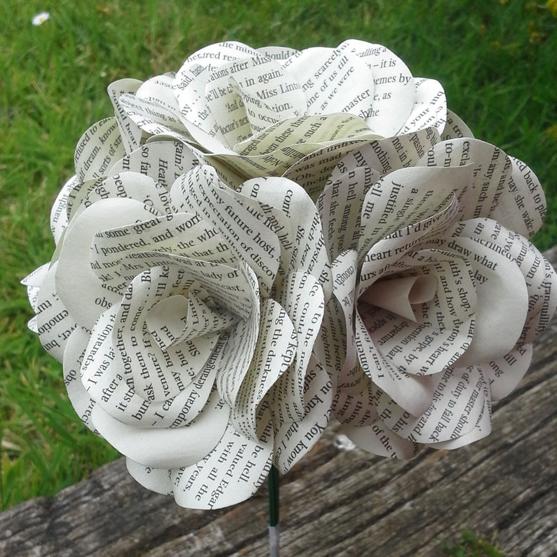 May include: A bouquet of paper flowers made from recycled book pages. The flowers are white and have a delicate, layered appearance. The bouquet is held together by a green stem.