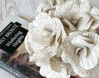 May include: A cluster of paper flowers crafted from book pages, featuring visible printed text, positioned next to a copy of the book Wuthering Heights by Emily Bronte. The flowers have layered, curled petals made from off-white paper.