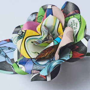 May include: A colourful paper flower crafted from comic book pages. The flower is composed of several layers of paper, each with a different pattern. The flower is a vibrant mix of red, yellow, blue, green, and black.