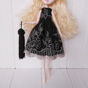 May include: A black tulle dress with silver embroidery and a scalloped lace trim. The dress is being worn by a doll with blonde hair and is holding a black tassel.